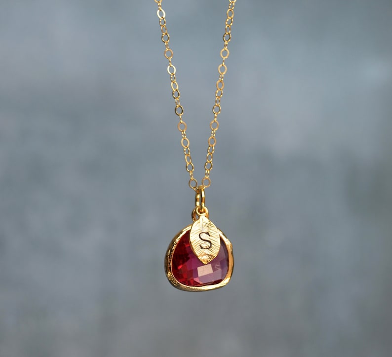 May include: Gold necklace with a red gemstone pendant and a small gold leaf charm with the letter 'S' engraved on it.