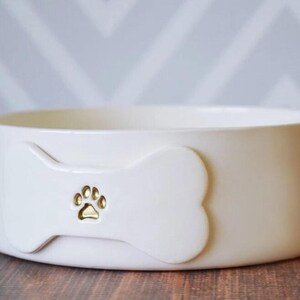 May include: A round, off-white ceramic pet bowl featuring a raised bone-shaped emblem on the front. The centre of the bone emblem displays a small, recessed gold-coloured paw print design. The bowl has a smooth, matte finish and a wide, open top.