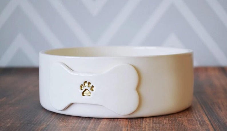 May include: A round, off-white ceramic pet bowl featuring a raised bone-shaped emblem on the front. The centre of the bone emblem displays a small, recessed gold-coloured paw print design. The bowl has a smooth, matte finish and a wide, open top.
