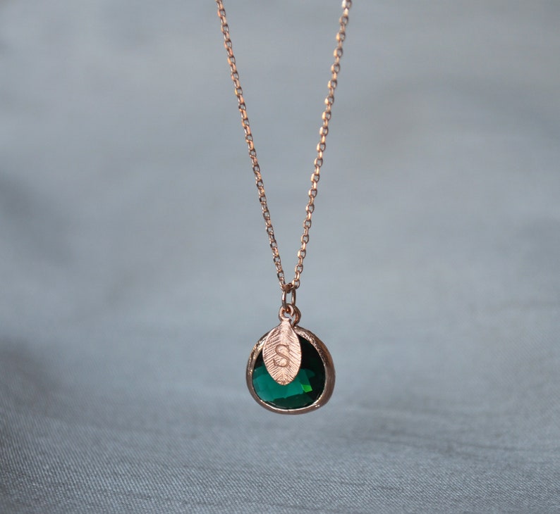 A green emerald colored transparent glass stone charm is framed in rose gold metal and hangs from a rose gold link chain. There is a small leaf shaped charm with the letter S stamped on the front, hanging in front of the emerald charm.