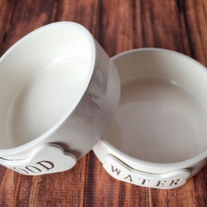 Two white ceramic pet bowls with bone-shaped name plates. The bowl on the left has the word "FOOD" on the handle and the bowl on the right has the word "WATER" on the handle.