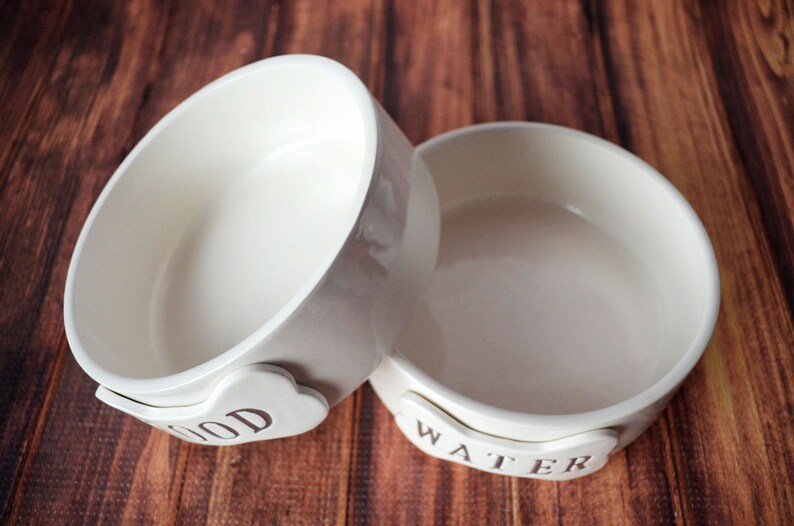Two white ceramic pet bowls with bone-shaped name plates. The bowl on the left has the word "FOOD" on the handle and the bowl on the right has the word "WATER" on the handle.