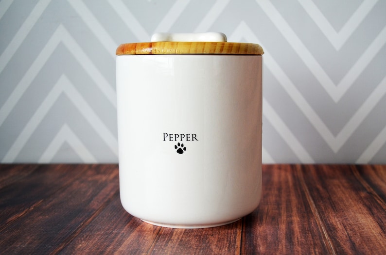 May include: White ceramic pet food container with a wooden lid. The container has the name "Pepper" and a paw print on the front.