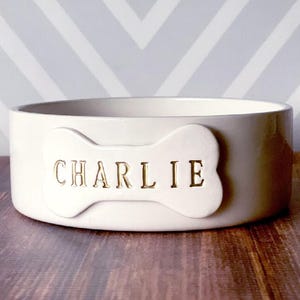 May include: A round, off-white ceramic pet bowl featuring a raised bone-shaped emblem on the front. The name CHARLIE is embossed in gold-coloured capital letters across the centre of the bone-shaped detail on the side of the bowl.