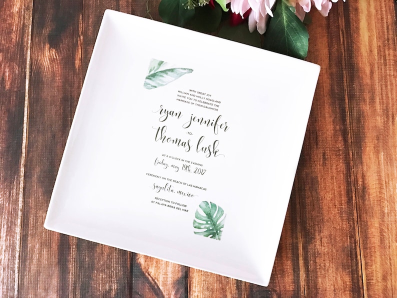 White ceramic square plate with a wedding invitation printed on it. The invitation features a tropical leaf design and the text "With great joy, William Holly Holland and Mary Elizabeth Graham invite you to celebrate the marriage of their daughter Ryan Jennifer to Thomas Lush at 4 o'clock in the evening Friday, May 19th, 2017 Ceremony on the beach of Las Palmas Sayulita, Mexico Reception to follow at Palapa Azul del Mar"
