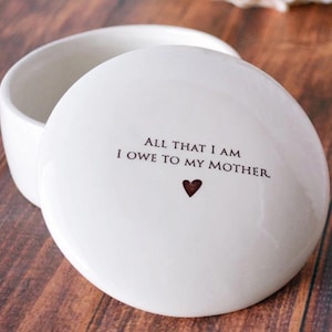 May include: White ceramic trinket box with a lid. The lid has the text "All that I am I owe to my mother." with a small red heart.
