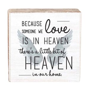 May include: A white wooden sign with a gray watercolor angel wing design. The sign reads "Because someone we love is in heaven, there's a little bit of heaven in our home."