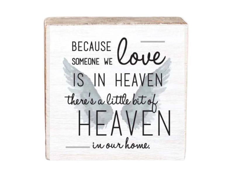 May include: A white wooden sign with a gray watercolor angel wing design. The sign reads "Because someone we love is in heaven, there's a little bit of heaven in our home."