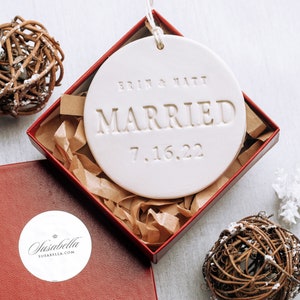 White ceramic ornament with the text "Erin & Matt Married 7.16.22" in a red box with brown tissue paper.