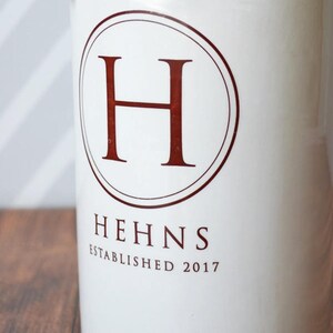 Puede incluir: Contenedor de cer&aacute;mica blanca con un emblema circular con la letra "H" en un tono rojo oscuro. La palabra "HEHNS" y el a&ntilde;o "ESTABLISHED 2017" est&aacute;n impresos debajo en el mismo color.