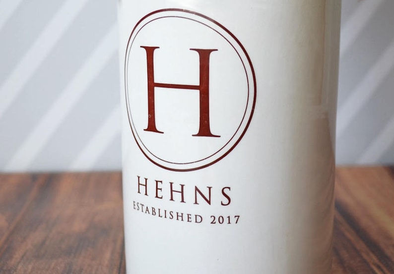 Puede incluir: Contenedor de cer&aacute;mica blanca con un emblema circular con la letra "H" en un tono rojo oscuro. La palabra "HEHNS" y el a&ntilde;o "ESTABLISHED 2017" est&aacute;n impresos debajo en el mismo color.
