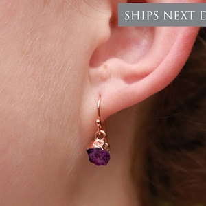 Op de afbeelding: Oorbellen van roségoud met een paarse edelsteen hanger. De oorbellen worden getoond op het oor van een persoon.