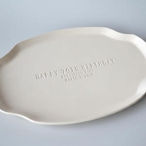 A handmade white ceramic serving platter with a gently scalloped edge. The platter is inscribed with "HAPPY 70TH BIRTHDAY" and the name "Mary Beth Ping" and the date "March 1, 2026". Plain background.