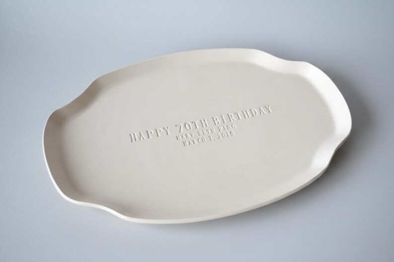 A handmade white ceramic serving platter with a gently scalloped edge. The platter is inscribed with "HAPPY 70TH BIRTHDAY" and the name "Mary Beth Ping" and the date "March 1, 2026". Plain background.