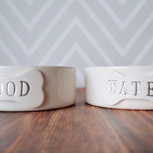 Two white ceramic dog bowls with bone-shaped tiles. The bowl on the left says "FOOD" and the bowl on the right says "WATER".