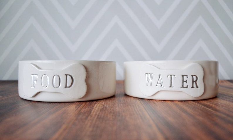 Two white ceramic dog bowls with bone-shaped tiles. The bowl on the left says "FOOD" and the bowl on the right says "WATER".