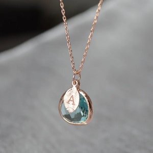 A light blue aquamarine colored transparent glass stone charm is framed in rose gold metal and hangs from a rose gold link chain. There is a small leaf shaped charm with the letter A stamped on the front, hanging in front of the aquamarine charm.