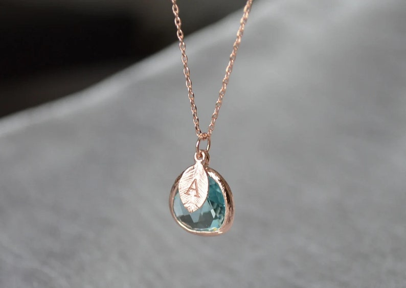 A light blue aquamarine colored transparent glass stone charm is framed in rose gold metal and hangs from a rose gold link chain. There is a small leaf shaped charm with the letter A stamped on the front, hanging in front of the aquamarine charm.