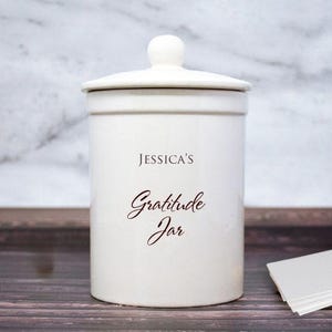 May include: A white ceramic mug with a black handle and the text "But First, Coffee" in a bold, black font. The mug is sitting on a wooden surface. The mug is approximately 4 inches tall.