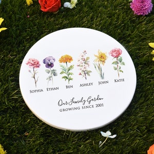 Puede incluir: Un azulejo de cerámica blanco redondo con un diseño floral. El azulejo presenta seis flores diferentes con nombres escritos debajo de cada flor: Sophia, Ethan, Ben, Ashley, John y Katie. El texto "Our Family Garden Growing Since 2001" está escrito debajo de las flores.