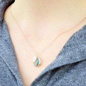 The necklace is shown on a model wearing a grey button down shirt. A rose gold metal chain with aquamarine charm and leaf charm hangs around the model's neck. The aquamarine is framed in rose gold and the leaf on top is stamped with an initial.