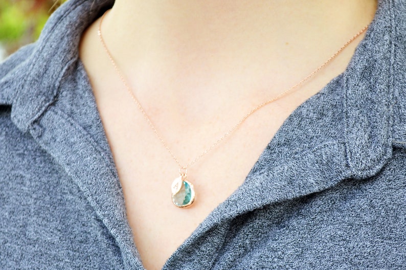 The necklace is shown on a model wearing a grey button down shirt. A rose gold metal chain with aquamarine charm and leaf charm hangs around the model's neck. The aquamarine is framed in rose gold and the leaf on top is stamped with an initial.