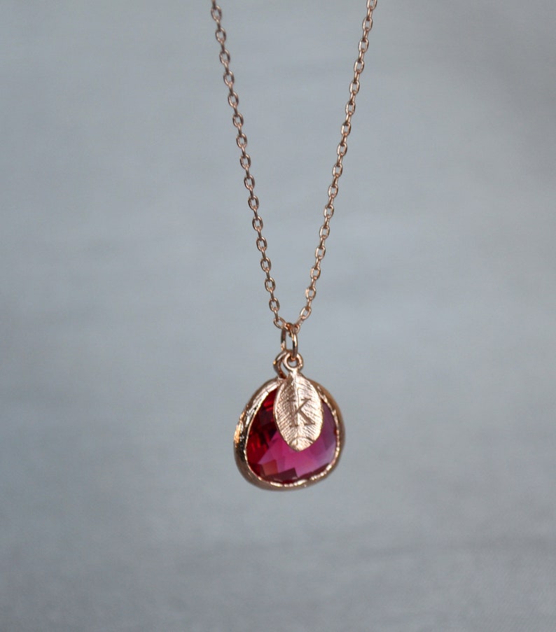 A pinkish red ruby colored transparent glass stone charm is framed in rose gold metal and hangs from a rose gold link chain. There is a small leaf shaped charm with the letter K stamped on the front, hanging in front of the ruby charm.