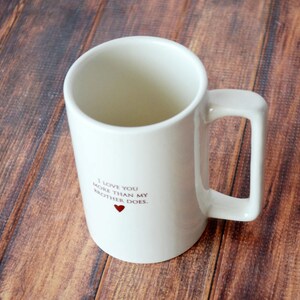 May include: White ceramic mug with a black inscription that reads "I love you more than my brother does." A small red heart is printed below the text.