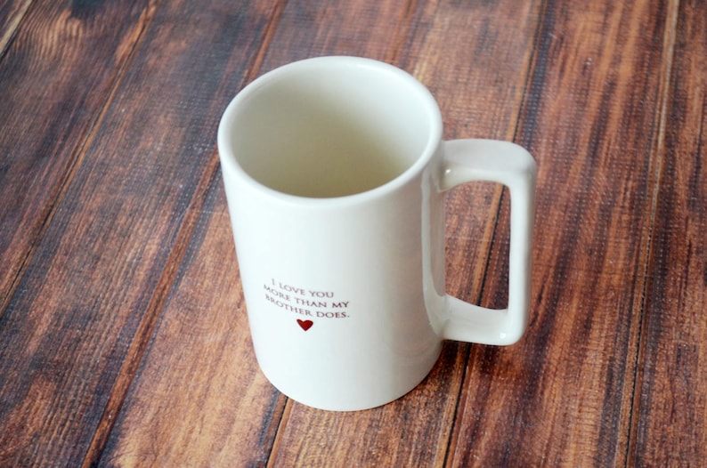 May include: White ceramic mug with a black inscription that reads "I love you more than my brother does." A small red heart is printed below the text.