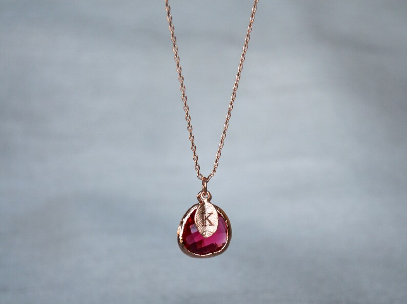 A pinkish red ruby colored transparent glass stone charm is framed in rose gold metal and hangs from a rose gold link chain. There is a small leaf shaped charm with the letter K stamped on the front, hanging in front of the ruby charm.