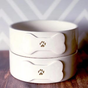 May include: Two stacked off-white ceramic bowls with a smooth, glossy finish. Each bowl features a raised, bone-shaped emblem on the front, centred with a small, debossed paw print design. The bowls have a simple, cylindrical shape.