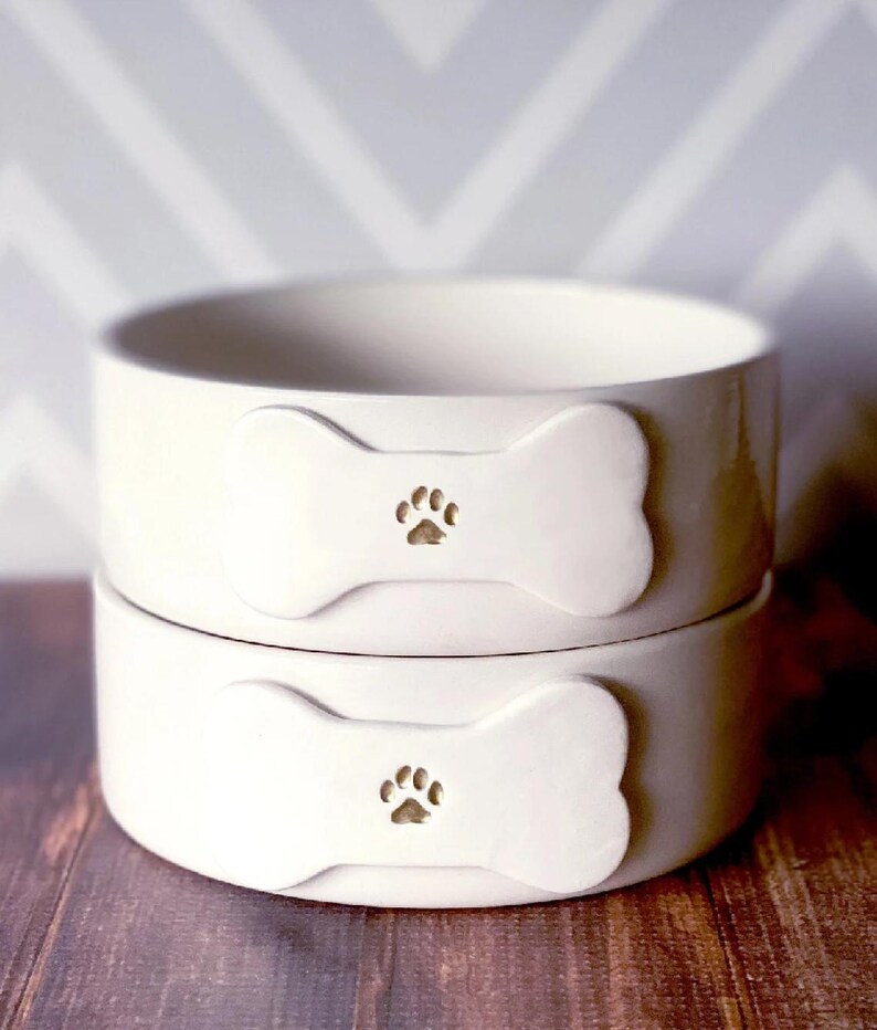 May include: Two stacked off-white ceramic bowls with a smooth, glossy finish. Each bowl features a raised, bone-shaped emblem on the front, centred with a small, debossed paw print design. The bowls have a simple, cylindrical shape.
