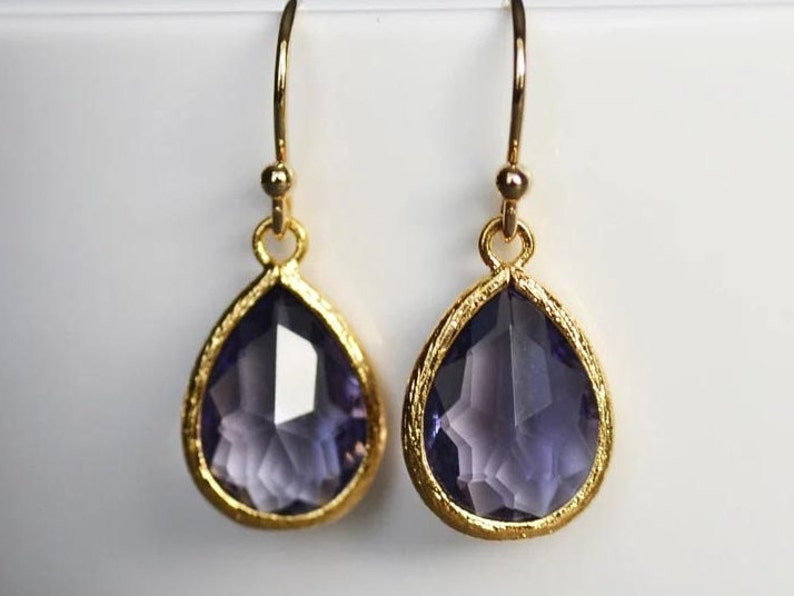 K&ouml;nnte beinhalten: Ein Paar goldene Tropfenohrringe mit violetten facettierten Glassteinen.