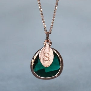 A green emerald colored transparent glass stone charm is framed in rose gold metal and hangs from a rose gold link chain. There is a small leaf shaped charm with the letter S stamped on the front, hanging in front of the emerald charm.