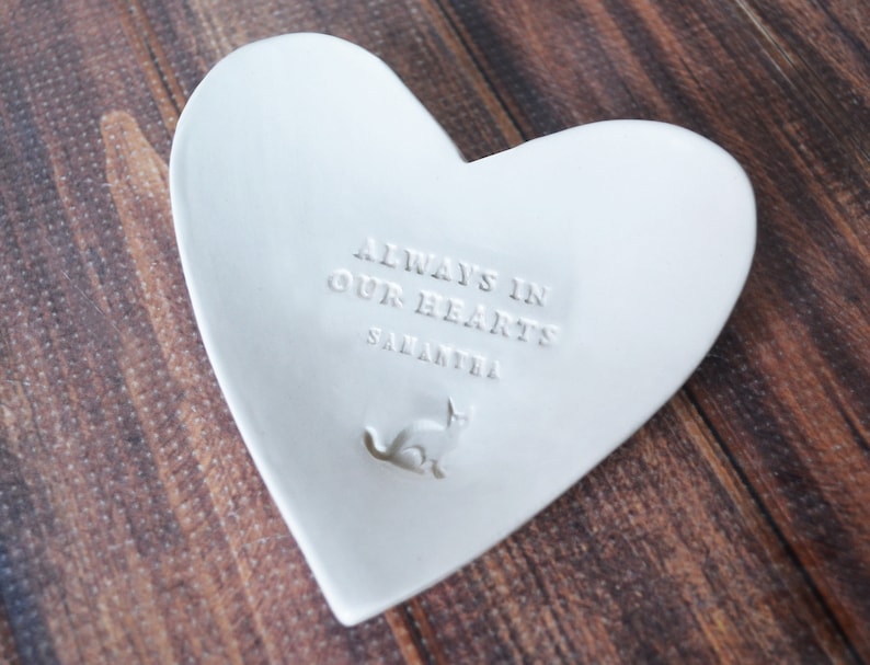 May include: White ceramic heart-shaped dish with the inscription "Always in our hearts" and the name "Samantha" below. A small cat silhouette is embossed in the center.