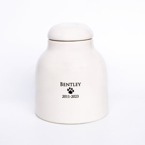 Pode incluir: Urna de cer&acirc;mica branca para animais de estima&ccedil;&atilde;o com uma pegada de pata preta e o nome "Bentley" e as datas "2011-2023" inscritas na frente.