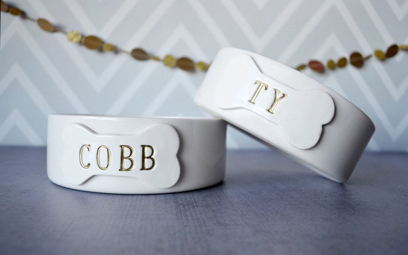 May include: Two white ceramic pet bowls with a smooth, glossy finish. Each bowl features a raised bone-shaped plaque on the front. The left bowl has the name COBB in gold lettering, and the right bowl has the name TY in gold lettering.