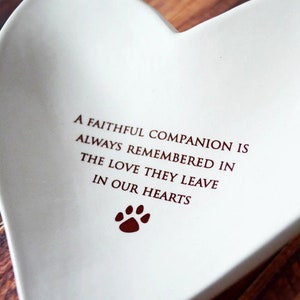 May include: White ceramic heart-shaped dish with a red paw print and the inscription "A faithful companion is always remembered in the love they leave in our hearts."
