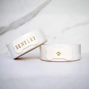 May include: Two white ceramic pet bowls featuring a bone-shaped emblem on the front. One bowl displays the name BENTLEY in gold lettering, while the other bowl features a gold paw print icon centred on the white bone-shaped detail.
