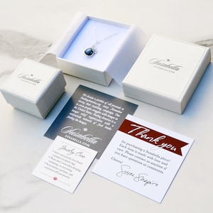 Puede incluir: Una caja de regalo blanca con un collar de plata y un colgante de piedra preciosa azul oscuro en su interior. La caja est&aacute; abierta y el collar es visible. La caja tiene el texto "Jasabella SUSABELLA.COM" impreso en ella. Tambi&eacute;n se puede ver una tarjeta de agradecimiento con el texto "&iexcl;Gracias por comprar una pieza de Susabella! Cada art&iacute;culo est&aacute; hecho con amor y cuidado. Por favor, contacta con nosotros en cualquier momento si tienes alguna pregunta o comentario. &iexcl;Saludos! Susan Shapiro".
