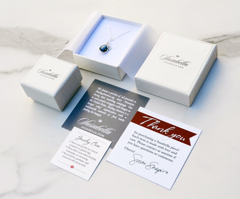 Informational image showing the included gift packaging for the necklace. Jewelry comes in a white gift box with Susabella logo, care directions, small business information, and thank you card. All package contents are displayed on a marble table.