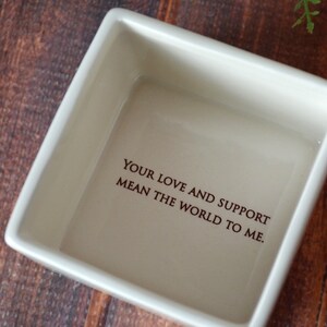 May include: A square, off-white ceramic dish with the inscription "YOUR LOVE AND SUPPORT MEAN THE WORLD TO ME" in dark brown lettering. The dish is set against a dark wood grain background.