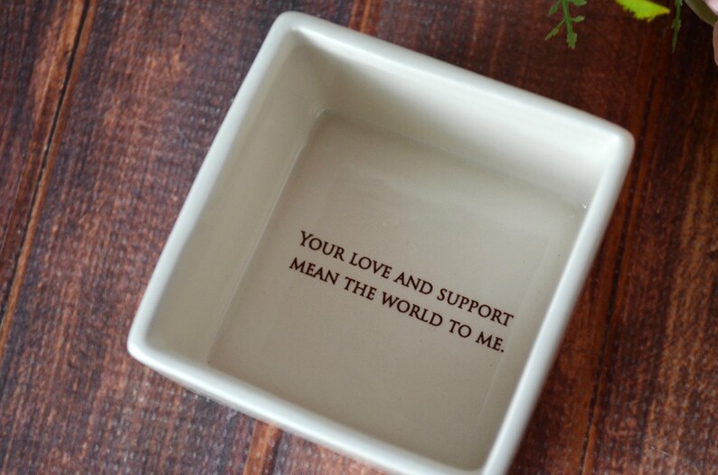 May include: A square, off-white ceramic dish with the inscription "YOUR LOVE AND SUPPORT MEAN THE WORLD TO ME" in dark brown lettering. The dish is set against a dark wood grain background.