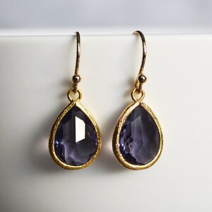 K&ouml;nnte beinhalten: Ein Paar goldene Tropfenohrringe mit violetten facettierten Steinen.