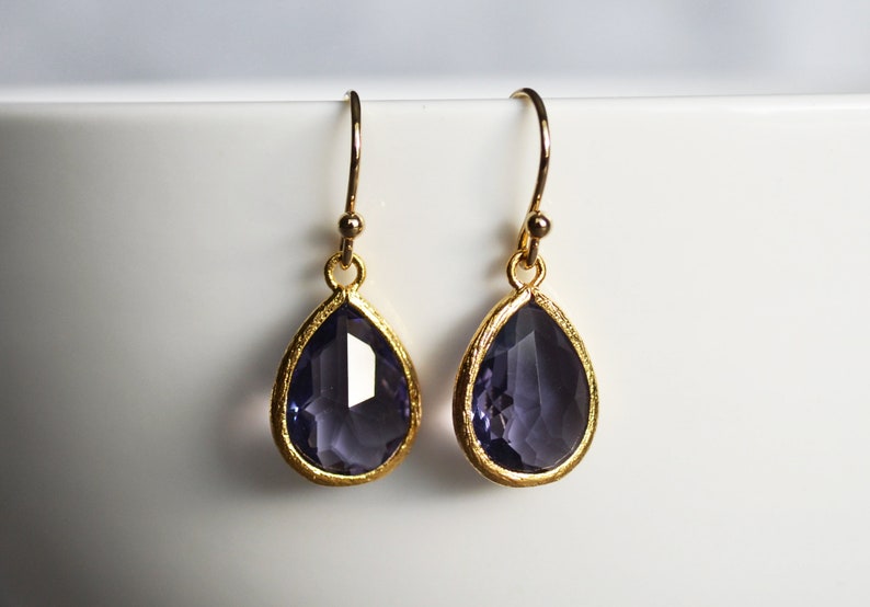 K&ouml;nnte beinhalten: Ein Paar goldene Tropfenohrringe mit violetten facettierten Steinen.