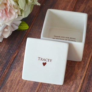 May include: White ceramic box with a lid. The lid has the name "TRACEY" and a small red heart. The inside of the box says "Thank you for being my Matron of Honor".