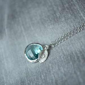 May include: A silver necklace with a round, light blue gemstone pendant and a leaf charm. The pendant is set in a silver bezel. The necklace chain is delicate and has a double strand design. The background is a soft, gray fabric.