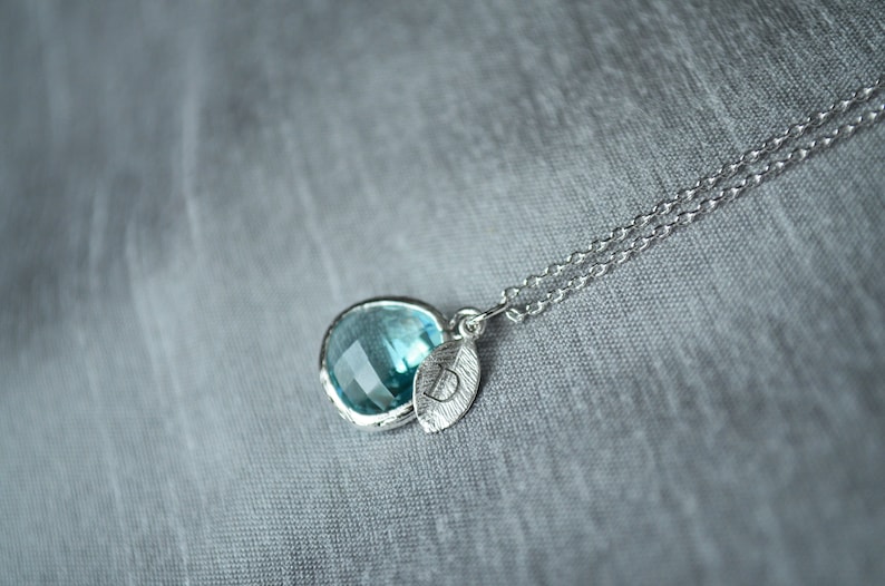 May include: A silver necklace with a round, light blue gemstone pendant and a leaf charm. The pendant is set in a silver bezel. The necklace chain is delicate and has a double strand design. The background is a soft, gray fabric.