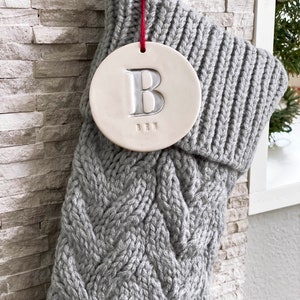 A grey knitted holiday stocking with off-white, round, ceramic ornament hanging from red cord. A large letter B is stamped into the ornament and painted in metallic silver. The name "BEN" is stamped below the initial in smaller text.