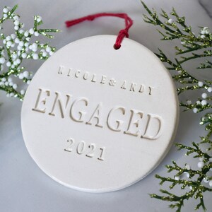 Product image of an off white gloss glazed ceramic round shaped  ornament. The front is stamped with custom text in all capital letters. The text is centered and reads Nicole & Andy, Engaged, 2021. The ornament has a red cord to hang from.